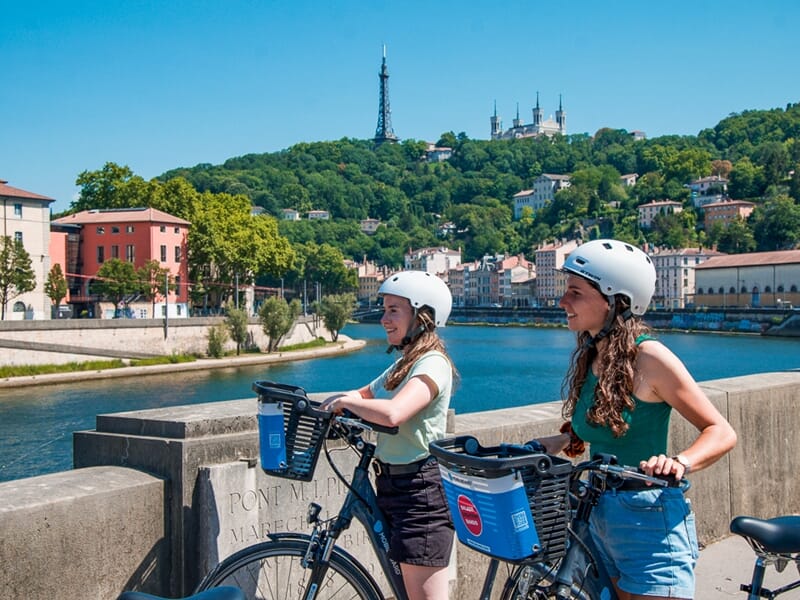 Deux jeunes femmes en balade vélo électrique Lyon, arrêt sur un pont avec vue sur la basilique de Fourvière et la colline verdoyante.