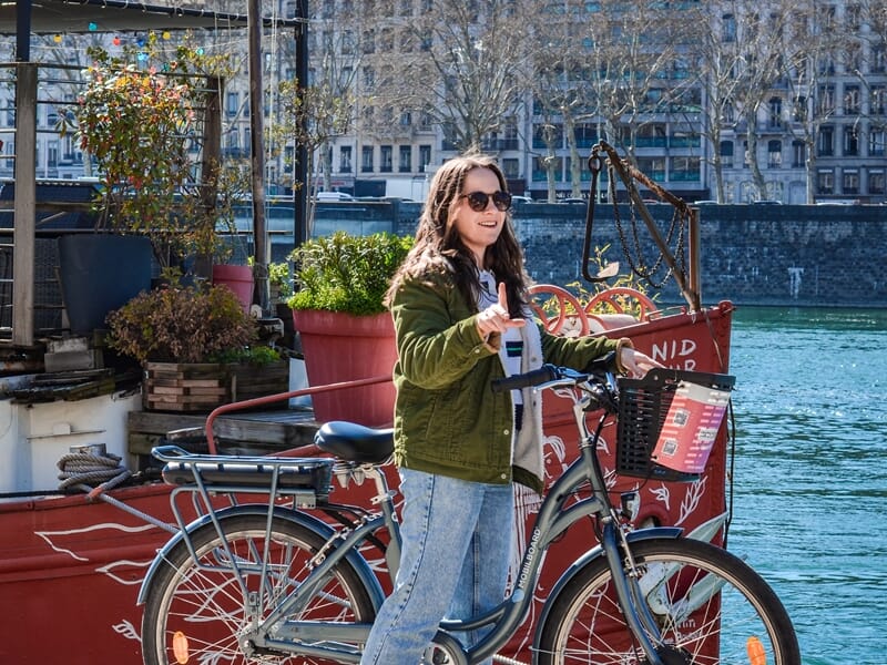 Balade vélo électrique Lyon sur les quais, une femme souriante avec son VAE stationné devant une péniche au bord du Rhône.