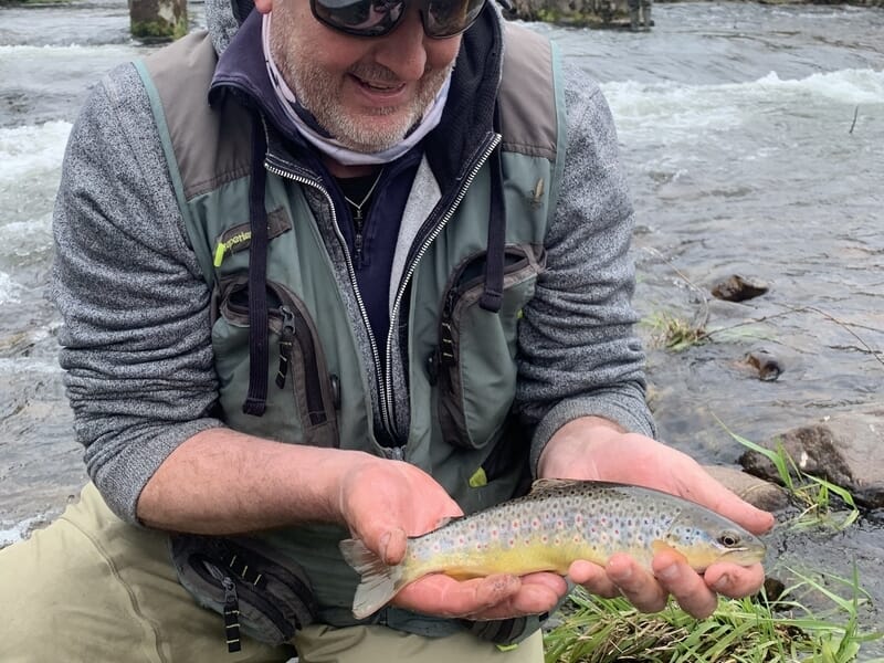 Homme souriant tenant une truite brune fraîchement pêchée au bord d'une rivière lors d'un stage de pêche à Schirmeck.