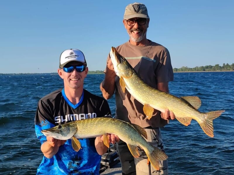 Deux hommes souriants tenant chacun un grand brochet pêché, sur un bateau lors d'un stage de pêche près de Strasbourg.
