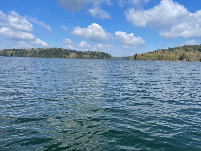 Vue panoramique du lac de Pierre-Percée avec une eau calme sous un ciel bleu parsemé de nuages blancs.