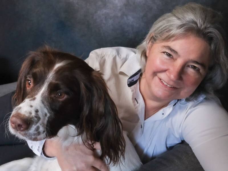 Une femme souriante en chemise blanche tient un grand chien blanc et marron, fond studio.