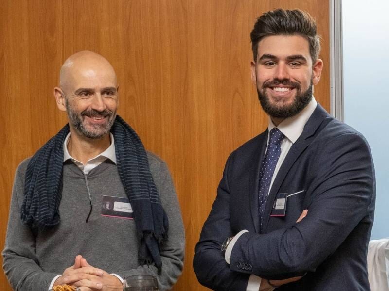 Deux hommes souriants en tenue professionnelle posent devant un mur en bois lors d'un événement formel.
