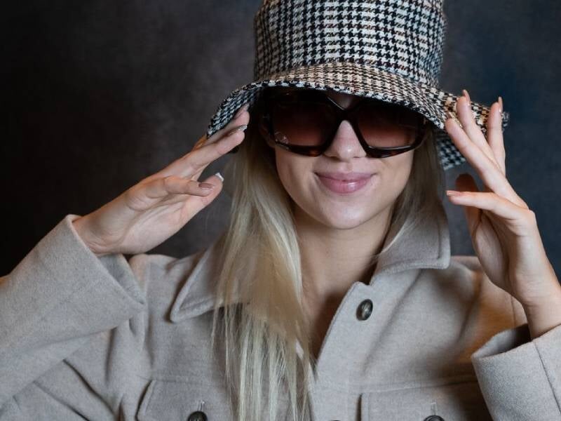 Jeune femme blonde portant un manteau beige, un chapeau à carreaux et de grandes lunettes de soleil noires.