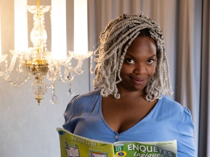 Femme aux cheveux tressés gris portant un haut bleu, tenant un magazine avec un lustre en arrière-plan.