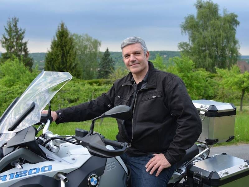 Homme mature en veste noire posant sur d'une moto BMW R1200 dans un cadre naturel verdoyant, près de Nanterre.