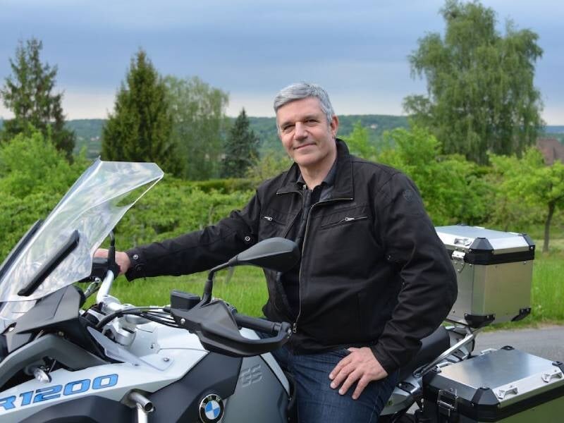 Homme mature en veste noire posant sur d'une moto BMW R1200 dans un cadre naturel verdoyant, près de Nanterre.