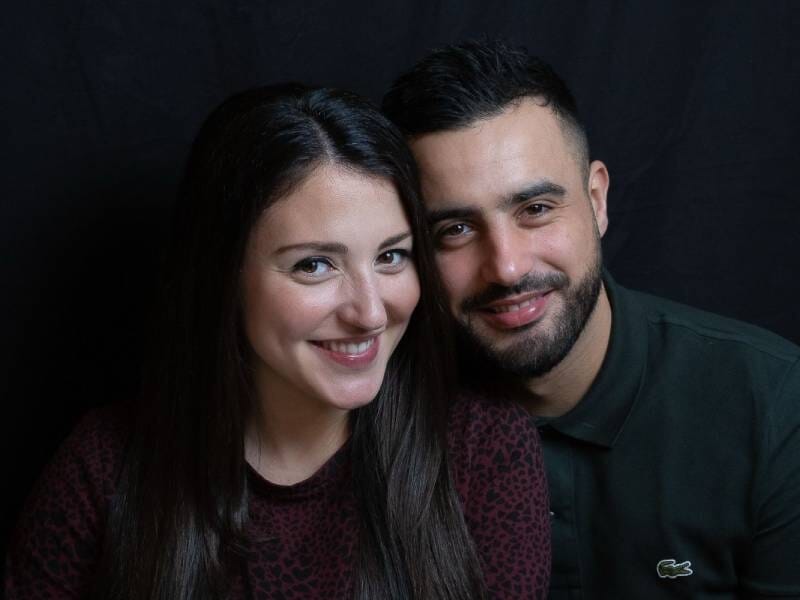 Portrait d'un couple souriant, une femme aux cheveux longs et un homme barbu, assis devant un fond noir uni, à Nanterre.