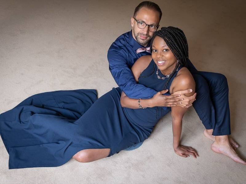 Un couple élégant pose ensemble sur un sol beige, la femme en robe bleue longue et l'homme en costume bleu marine.
