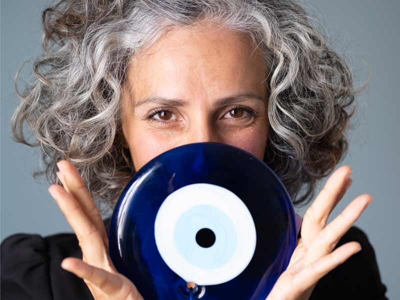 Femme aux cheveux gris bouclés tenant un disque bleu avec un motif œil, regardant directement l'appareil photo.