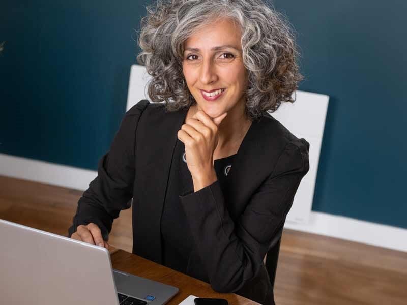 Femme souriante aux cheveux gris bouclés, assise à un bureau avec un ordinateur portable, posant la main sur le menton.