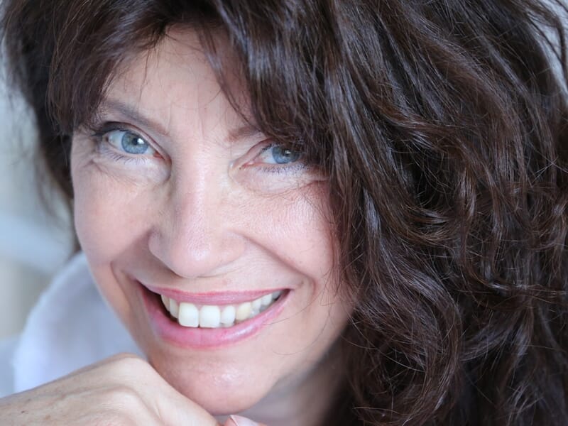Portrait d'une femme souriante aux cheveux bruns bouclés et aux yeux bleus, posant avec la main sous le menton.