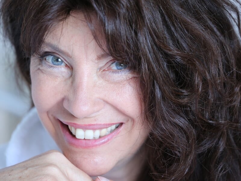 Portrait d'une femme souriante aux cheveux bruns bouclés et aux yeux bleus, posant avec la main sous le menton.