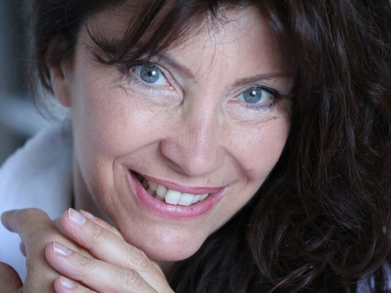 Portrait rapproché d'une femme aux cheveux bruns bouclés et yeux bleus, souriant avec les mains jointes devant elle.