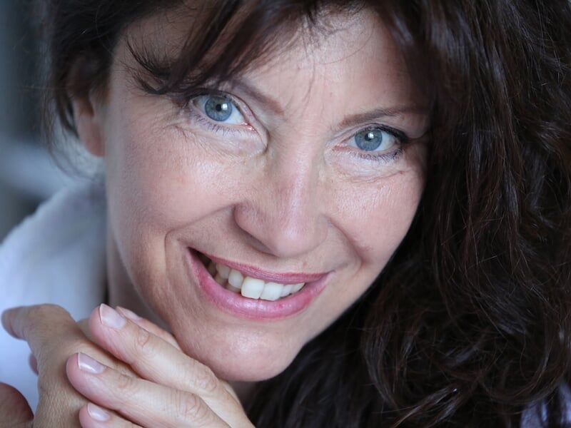 Portrait rapproché d'une femme aux cheveux bruns bouclés et yeux bleus, souriant avec les mains jointes devant elle.