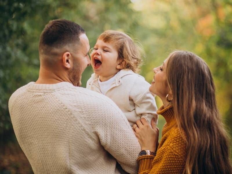 Un couple souriant tenant leur enfant qui rit, dans un cadre naturel verdoyant et ensoleillé.