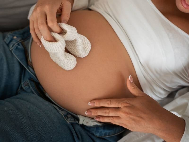 Femme enceinte allongée tenant une paire de chaussons blancs sur son ventre arrondi, en jean et t-shirt blanc.