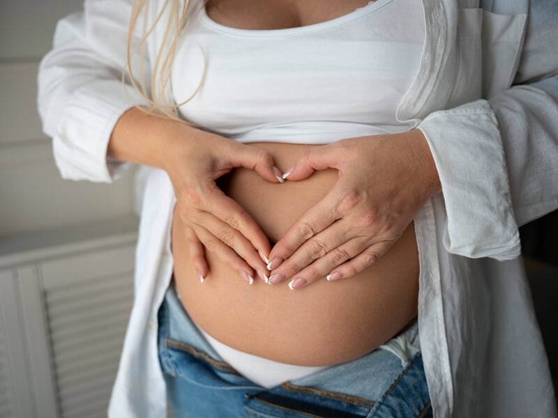 Couple formant un cœur avec leurs mains sur le ventre rond de la femme enceinte, habillée en blanc et jean.