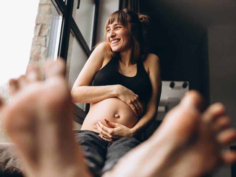 Femme enceinte souriante assise au sol, mains sur son ventre, pieds flous au premier plan, ambiance chaleureuse.