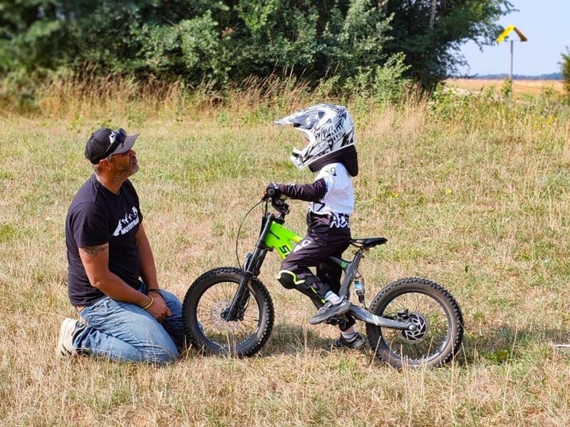 Un enfant en casque discute avec un adulte à genoux près d'une petite moto électrique dans un champ herbeux.