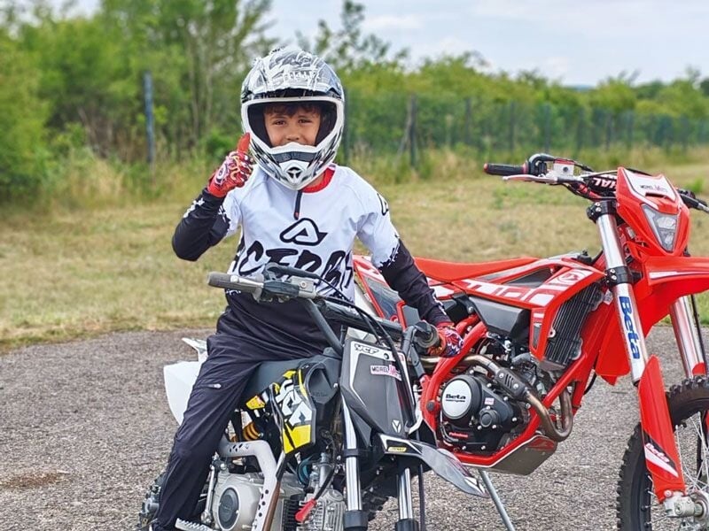 Un enfant en tenue de moto et casque fait un signe de la main assis sur une moto rouge et noire en extérieur.