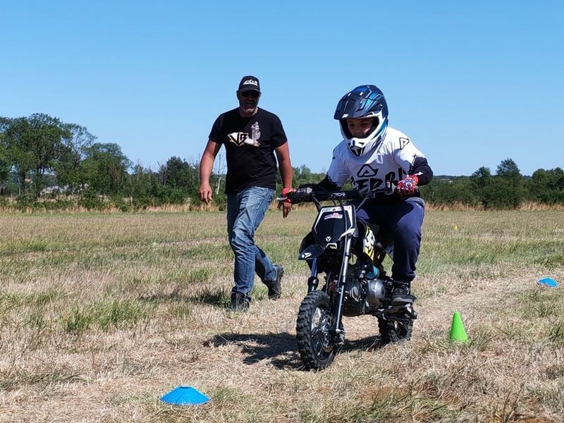 Un enfant en casque conduit une petite moto sur un terrain herbeux avec un adulte qui marche derrière lui.