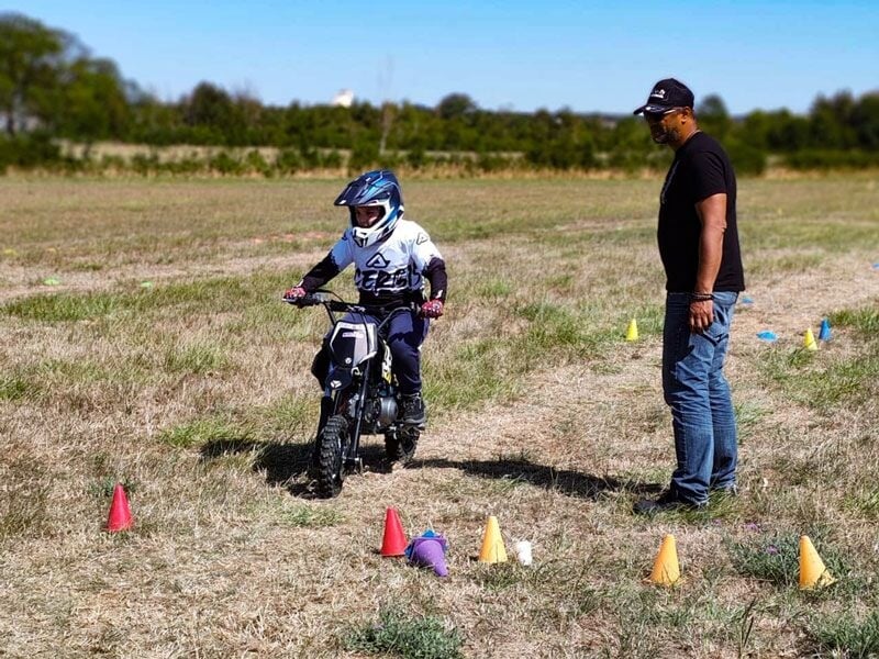 Un enfant en casque pilote une petite moto entre des cônes colorés sous la surveillance d'un adulte debout.