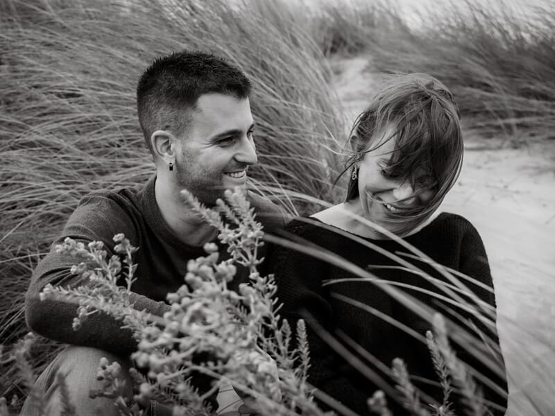 Un couple souriant assis dans les herbes hautes lors d'un shooting avec une photographe de couple à Béziers.
