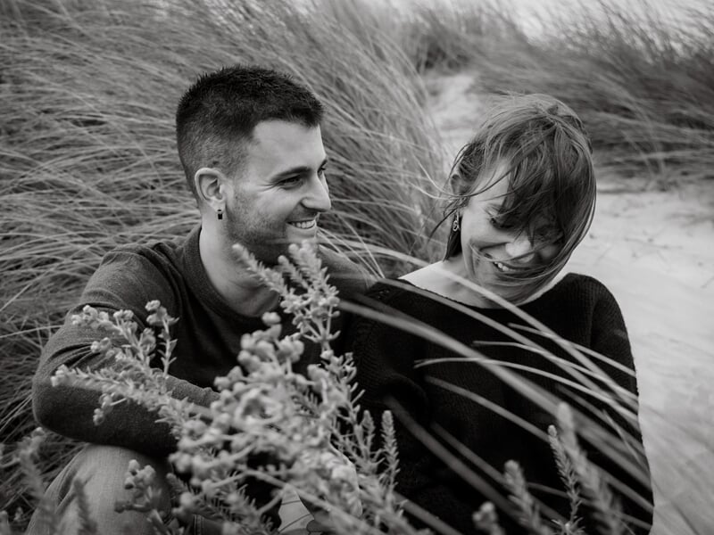 Un couple souriant assis dans les herbes hautes lors d'un shooting avec une photographe de couple à Béziers.