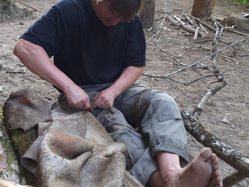 Homme assis par terre en forêt, travaillant avec un outil lors d'un stage de survie à Le Saint.