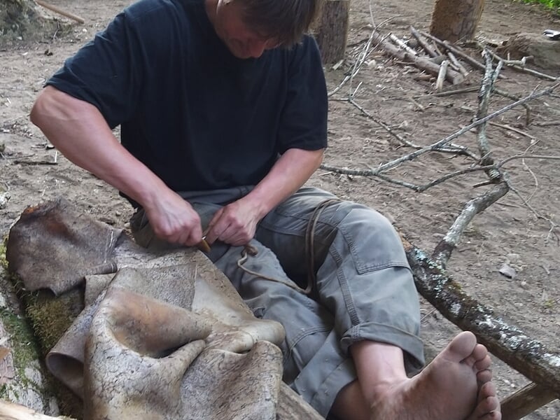 Homme assis par terre en forêt, travaillant avec un outil lors d'un stage de survie à Le Saint.