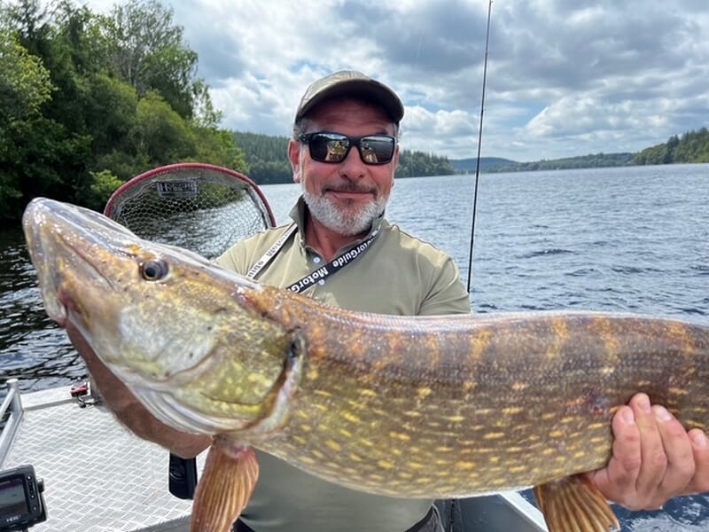 Homme souriant en bateau montrant un grand brochet fraîchement pêché lors d'un stage de pêche près de Limoges. 