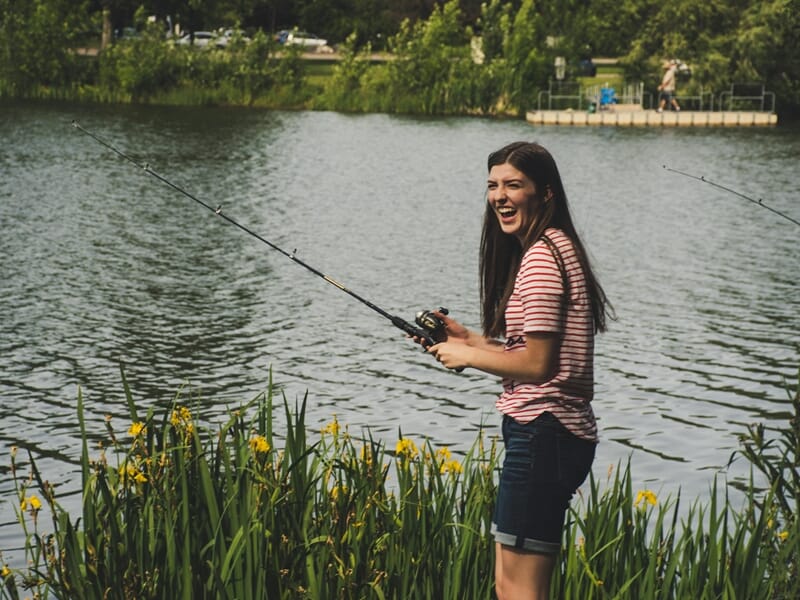 Jeune femme souriante en tenue décontractée pêchant au bord d'un lac lors d'un stage de pêche près d'Agen.