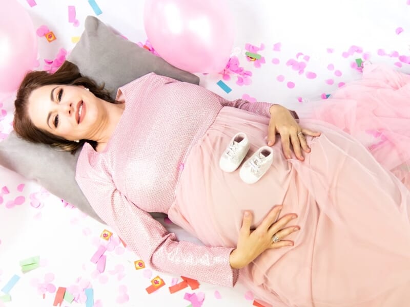 Femme enceinte allongée sur un coussin gris, entourée de ballons roses et confettis colorés, lors d'un shooting avec un photographe de grossesse à Nîmes.