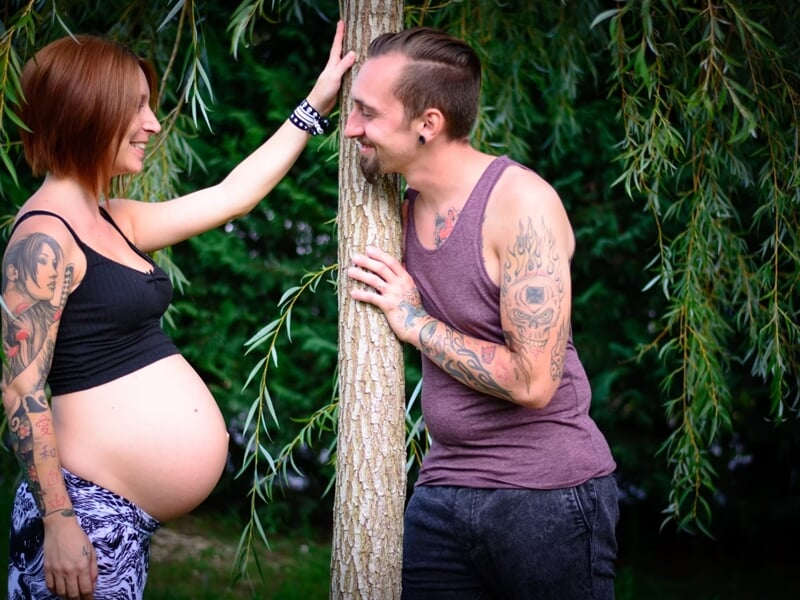 Couple tatoué souriant près d'un arbre, femme enceinte en débardeur noir lors d'un shooting avec un photographe de grossesse à Nîmes.