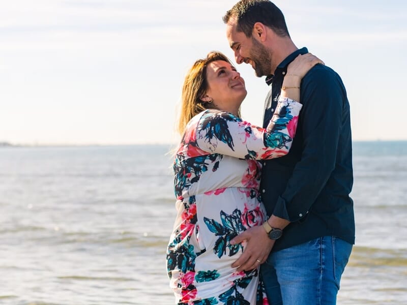 Couple souriant au bord de la mer, femme enceinte en robe fleurie, lors d'un shooting avec un photographe de grossesse à Nîmes.