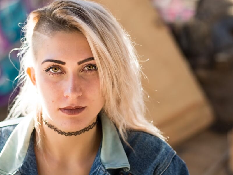Portrait d'une jeune femme blonde au regard intense, portant un collier noir et une veste en jean lors d'un shooting photo lifestyle à Nîmes.