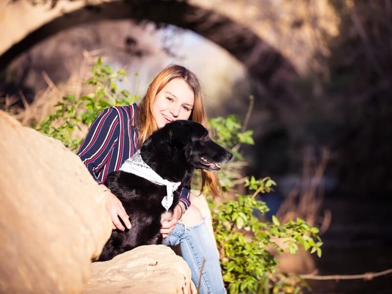 Femme souriante assise près d'un chien noir avec un foulard blanc, dans un cadre naturel ensoleillé lors d'un shooting photo lifestyle à Nîmes.