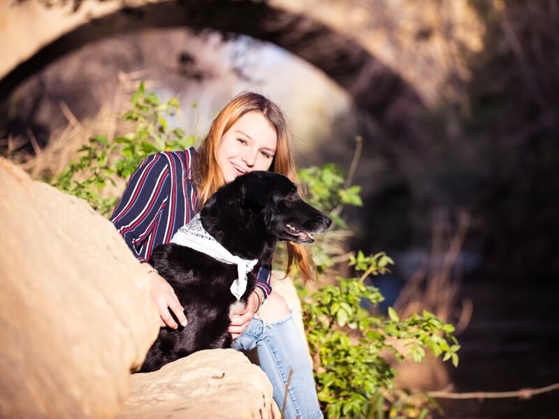 Femme souriante assise près d'un chien noir avec un foulard blanc, dans un cadre naturel ensoleillé lors d'un shooting photo lifestyle à Nîmes.