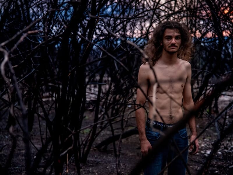 Homme torse nu avec cheveux longs, debout dans une forêt sombre lors d'un shooting photo lifestyle à Nîmes.