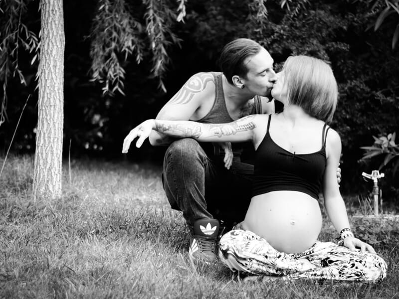 Un couple en extérieur, la femme enceinte assise sur l'herbe embrasse un homme lors d'un shooting photo couple à Nîmes.