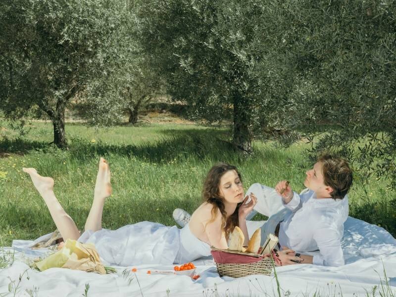 Un couple allongé sur une couverture blanche dans un champ d'oliviers, profitant d'un pique-nique ensoleillé.
