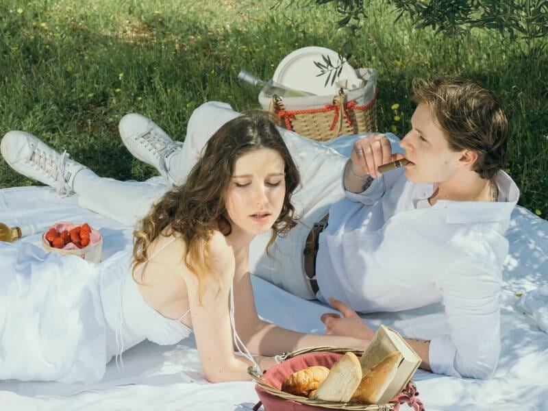Un couple en tenue blanche dégustant un pique-nique champêtre avec des fraises et du pain sous les arbres.