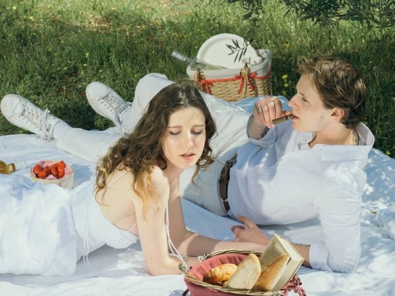 Un couple en tenue blanche dégustant un pique-nique champêtre avec des fraises et du pain sous les arbres.