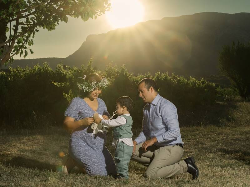 Famille en plein air, parents agenouillés avec leur enfant qui tient un jouet, sous un soleil éclatant.