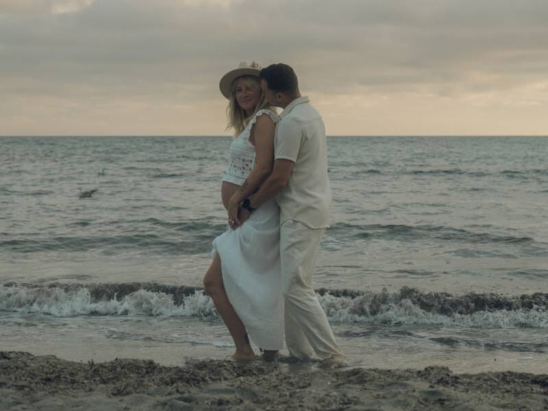 Un couple marche pieds nus sur la plage au bord de la mer sous un ciel nuageux au coucher du soleil.
