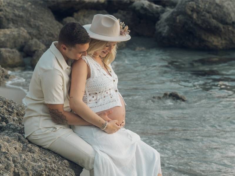 Un couple assis sur des rochers au bord de la mer, la femme enceinte porte un chapeau blanc, lors d'un shooting photo à Aix-en-Provence.