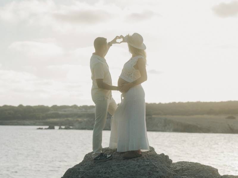 Un couple forme un cœur avec leurs mains devant le ventre de la femme enceinte, debout sur un rocher au bord de l'eau.
