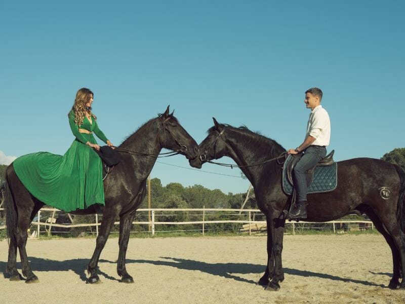 Un homme et une femme à cheval se font face dans une carrière, les chevaux noirs se touchent le museau.