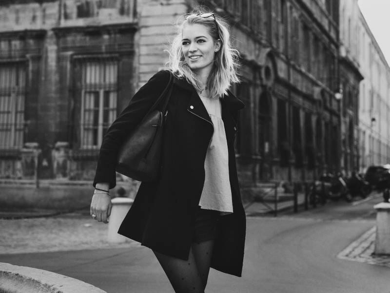 Jeune femme souriante en manteau noir marchant dans la rue, avec des bâtiments anciens en arrière-plan, lors d'un shooting photo lifestyle.